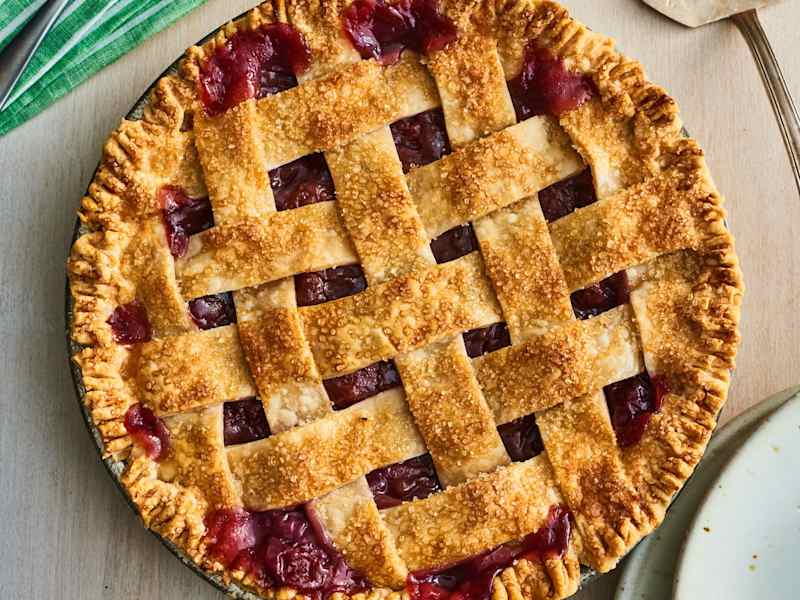 Lattice-top cherry pie with a golden crust, placed on a wooden surface next to a plate and utensils.