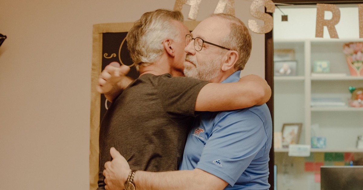 Two mature men embracing indoors, conveying friendship and affection.