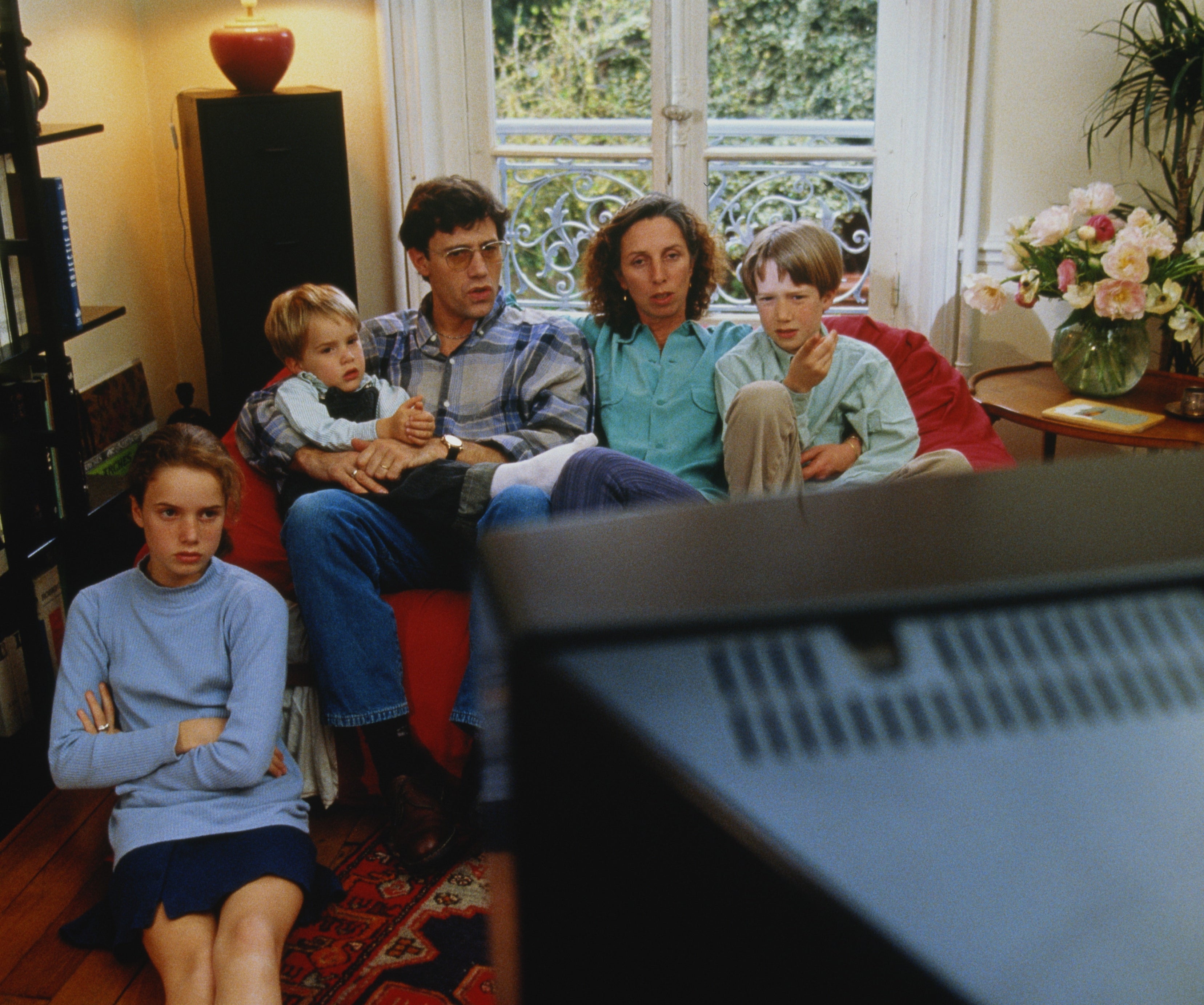 Family of five sitting on a couch in a living room, watching TV, with expressions of concentration