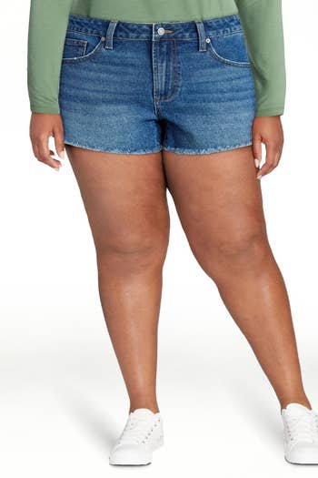 Person wearing denim shorts, green long-sleeve top, and white sneakers. Featured in shopping article