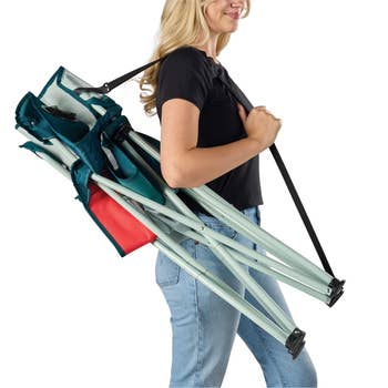 Person carrying a folded portable camping chair with a strap over their shoulder, wearing a casual black top and jeans