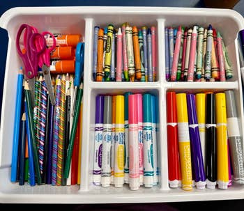 Organized tray with colorful craft supplies: scissors, glue sticks, pencils, crayons, and markers placed in separate compartments