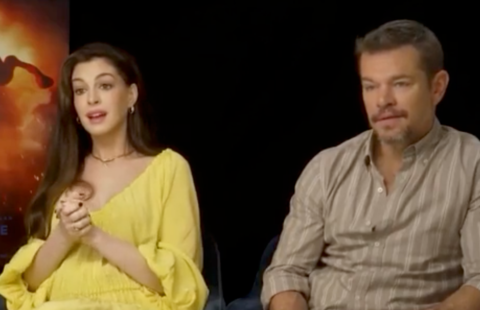 Two people seated in an interview setting. The woman wears a flowing dress, and the man is in a casual button-up shirt