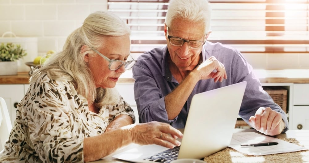 An elderly couple sits at a table, smiling and looking at a laptop, with papers and a pen nearby.