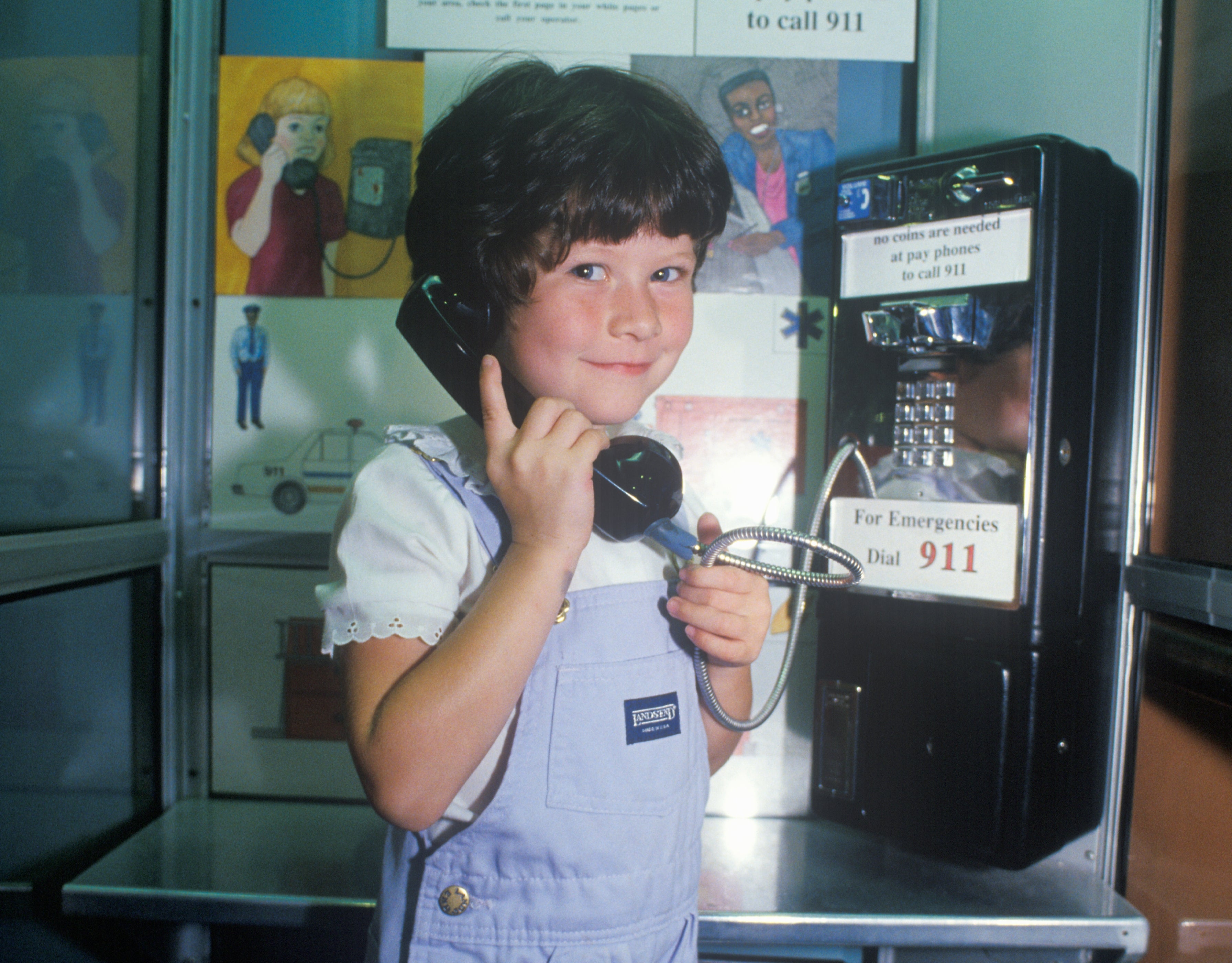 Child using a black rotary pay phone in a phone booth, smiling while holding the receiver to their ear. Vintage pay phone setting