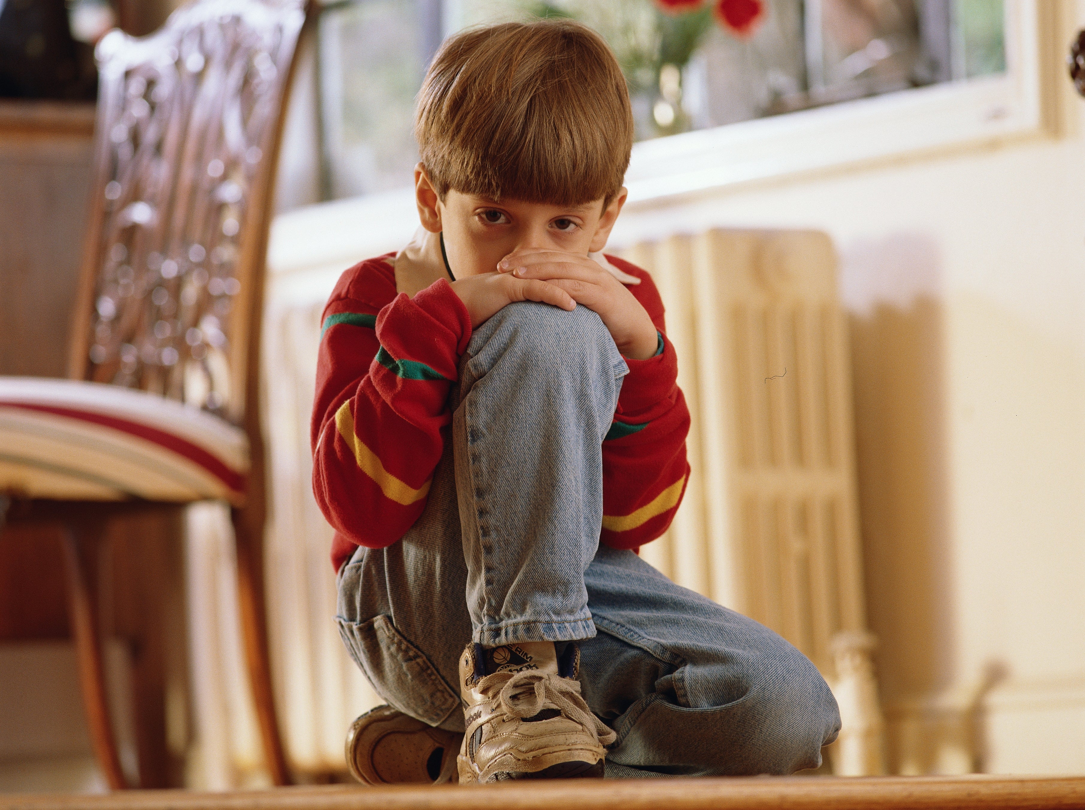 Child sitting indoors, wearing a long-sleeve top and jeans, looking thoughtful with chin on knees