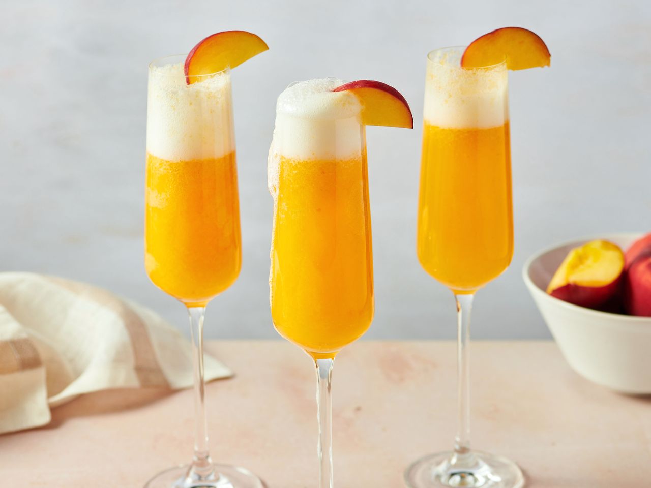 Three glasses of peach bellinis garnished with peach slices on a table with a napkin and a bowl of peaches