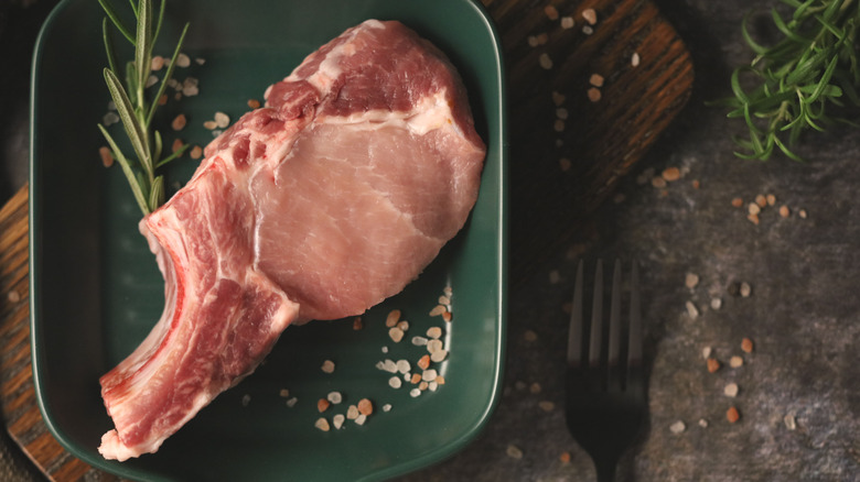 Raw bone-in and thick-cut pork chop on green square dish surrounded by fresh rosemary sprigs and salt crystals