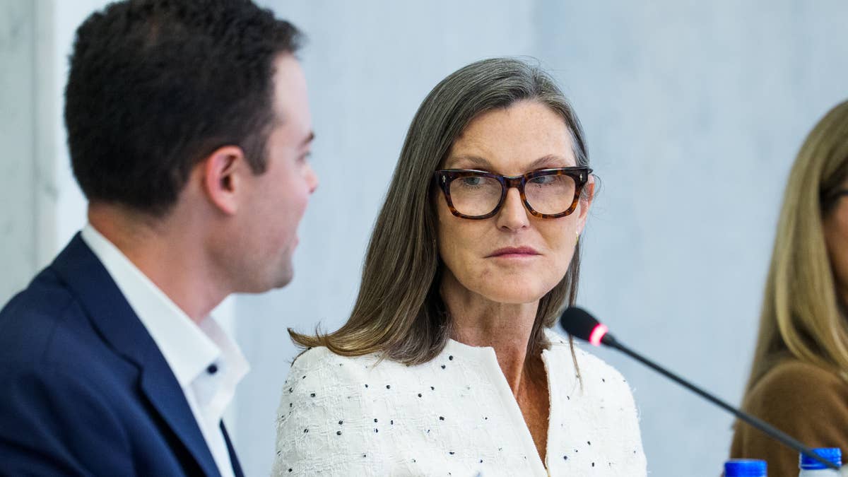ark-invest-ceo-cathie-wood-speaks-to-a-man-during-an-event-sitting-in-front-of-a-white-wall