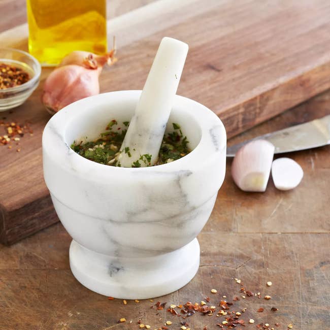 Marble mortar and pestle with herbs, placed on a wooden surface, next to shallots, chili flakes, knife, and olive oil bottle. Perfect for kitchen use