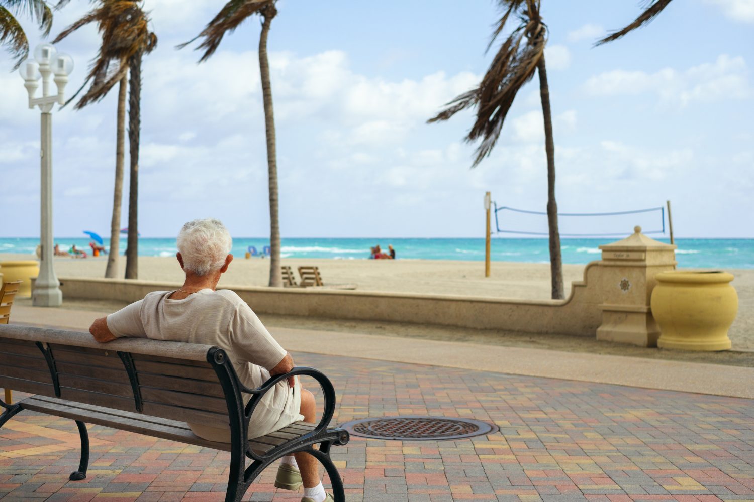 From behind, an elderly person with white hair, wearing a beige shirt and shorts, sits on a dark wooden and metal park bench. They are looking towards a sandy beach with turquoise ocean water and distant palm trees under a blue sky with scattered clouds. A volleyball net stands on the beach, and the foreground features a colorful brick walkway.