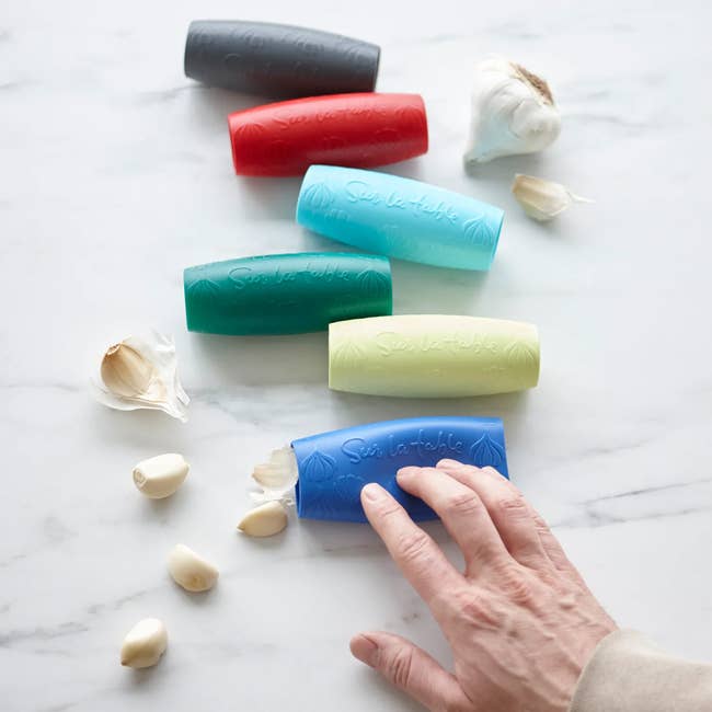 Silicone garlic peelers in various shades are scattered on a marble surface with garlic cloves nearby. A hand reaches for one peeler