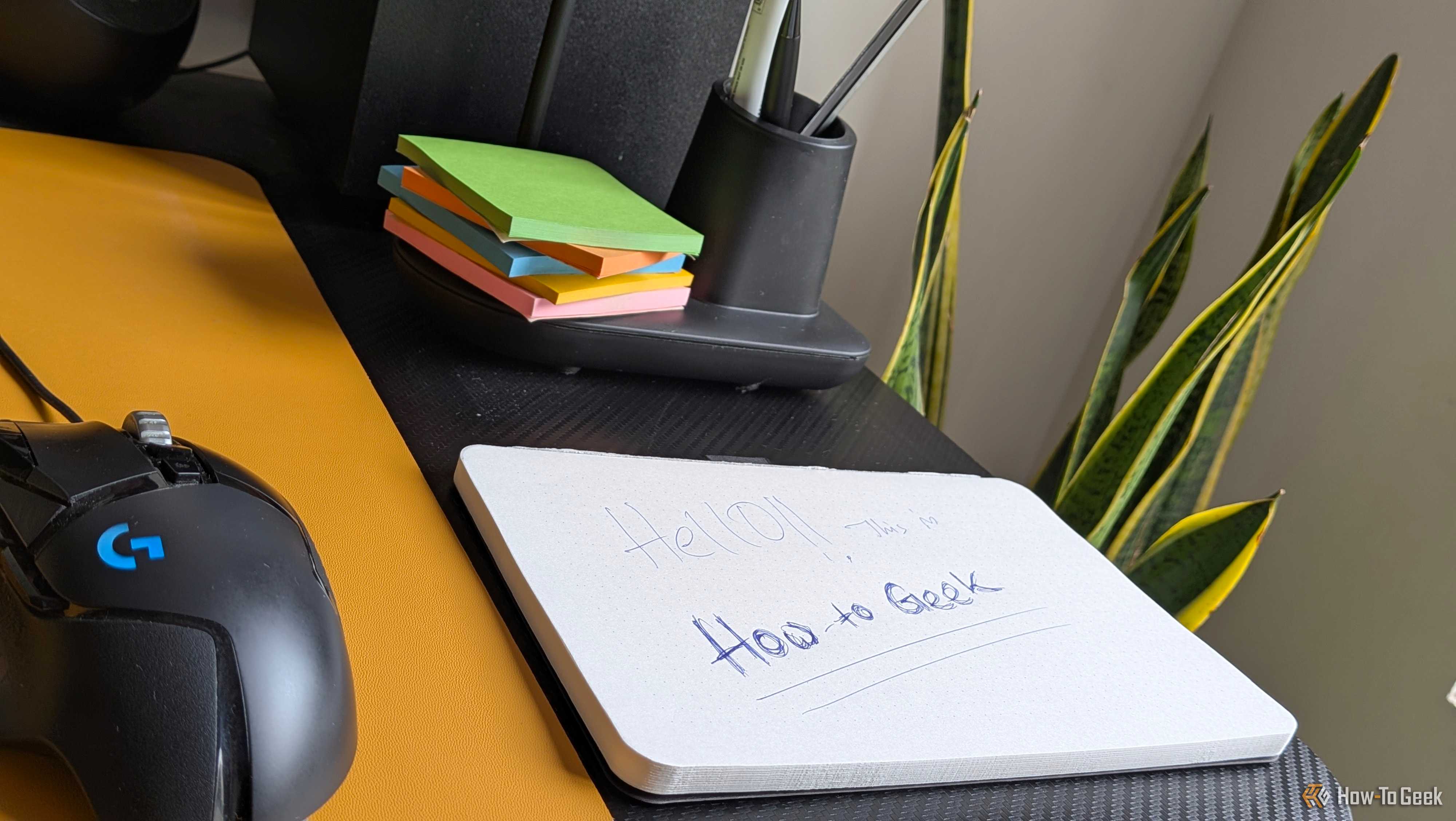 Notepad with How to Geek intro note resting on desk next to sticky notes and pen holder.