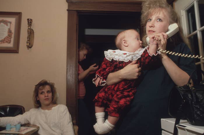 Woman holds a baby while talking on the phone; another woman sits at a table, and a third person is in the background holding a device