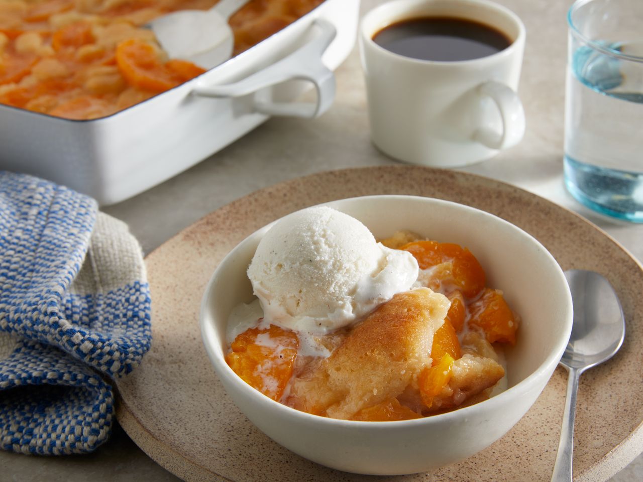 high view of peach cobbler, topped with melty vanilla ice cream and a cup of coffee in the background