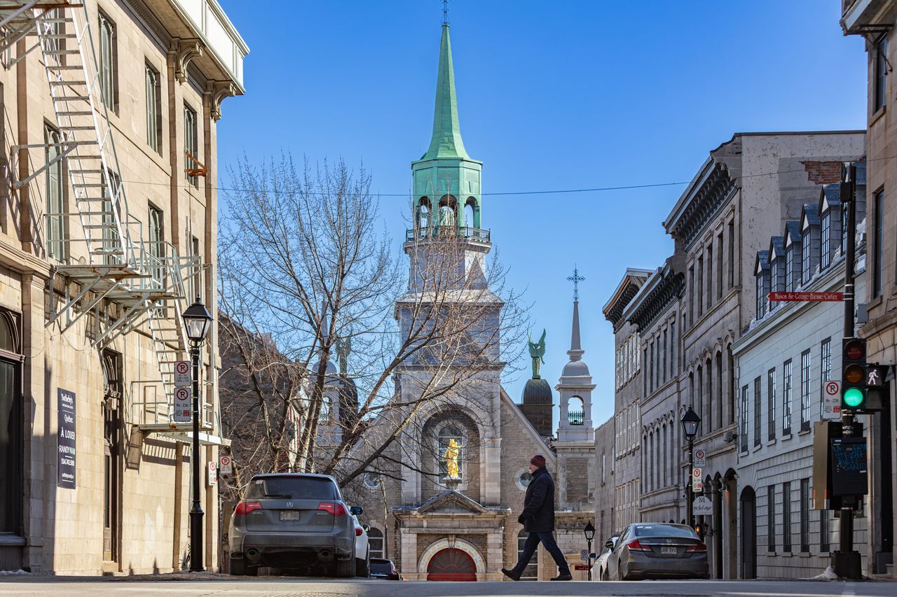 A person walks down a street with a church featuring a tall spire in the background