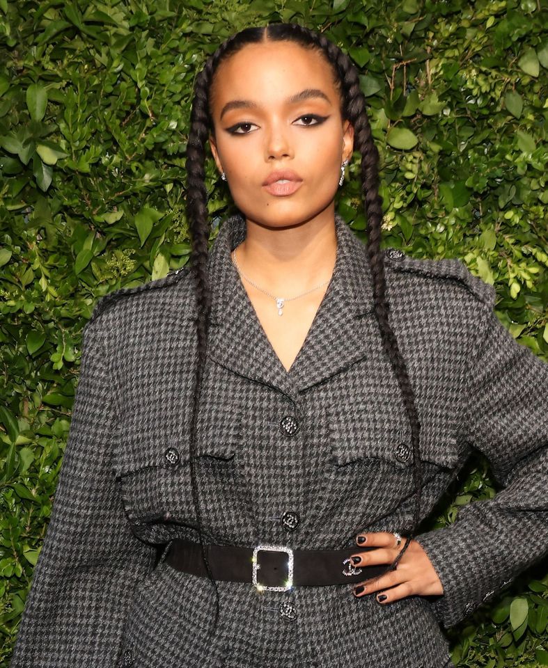 A woman with long braided hair standing in front of a leafy background wearing a belted checkered top