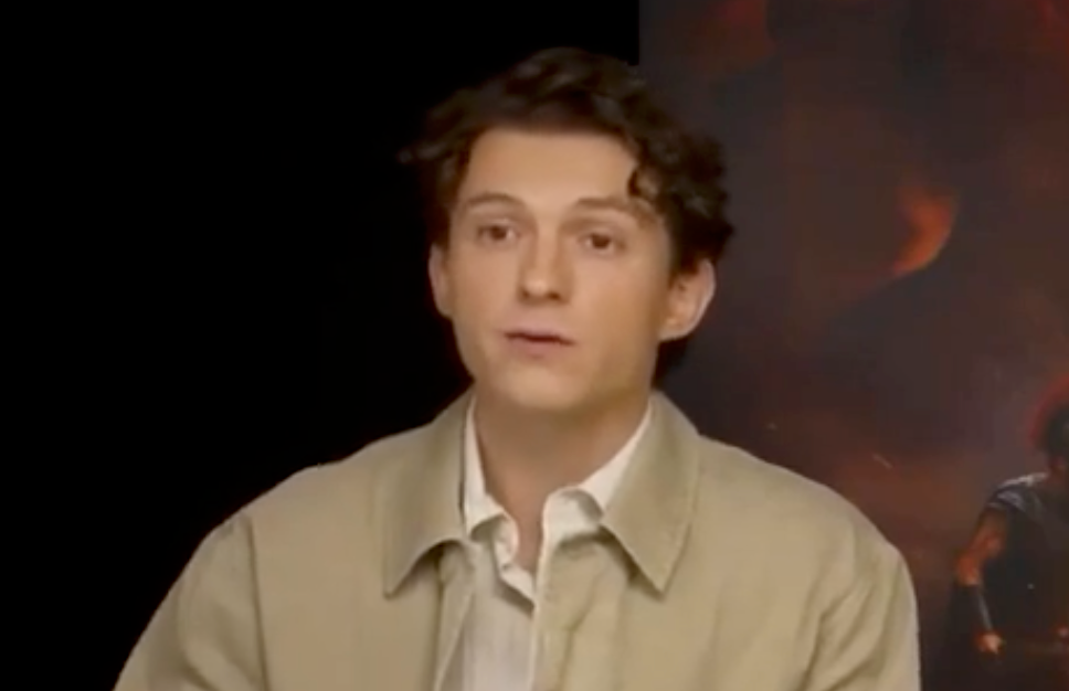 A person with curly hair wearing a light jacket over a button-up shirt sits in front of a blurred backdrop