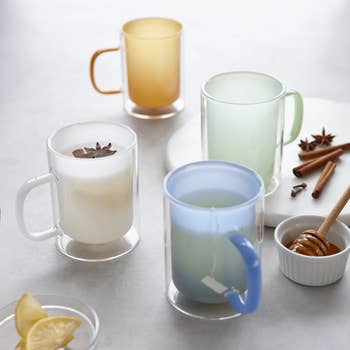 Four glass mugs with different teas and spices on a table, alongside lemon wedges and honey, creating a cozy beverage display