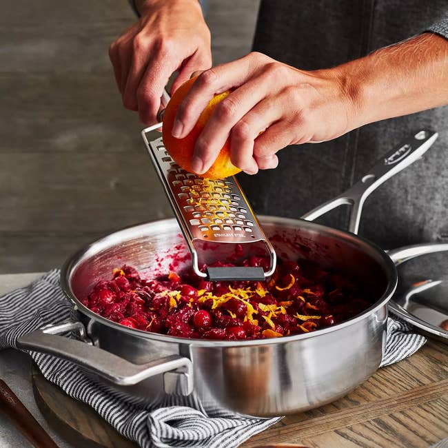 Person grating orange zest over a pan of food on a wooden board, suggesting a cooking or kitchen accessory available for purchase