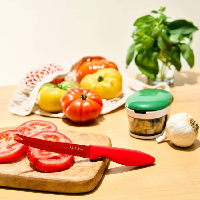 Sliced tomatoes on a wooden board with a red-handled knife, surrounded by whole tomatoes, basil, garlic, and a vegetable chopper on a kitchen counter