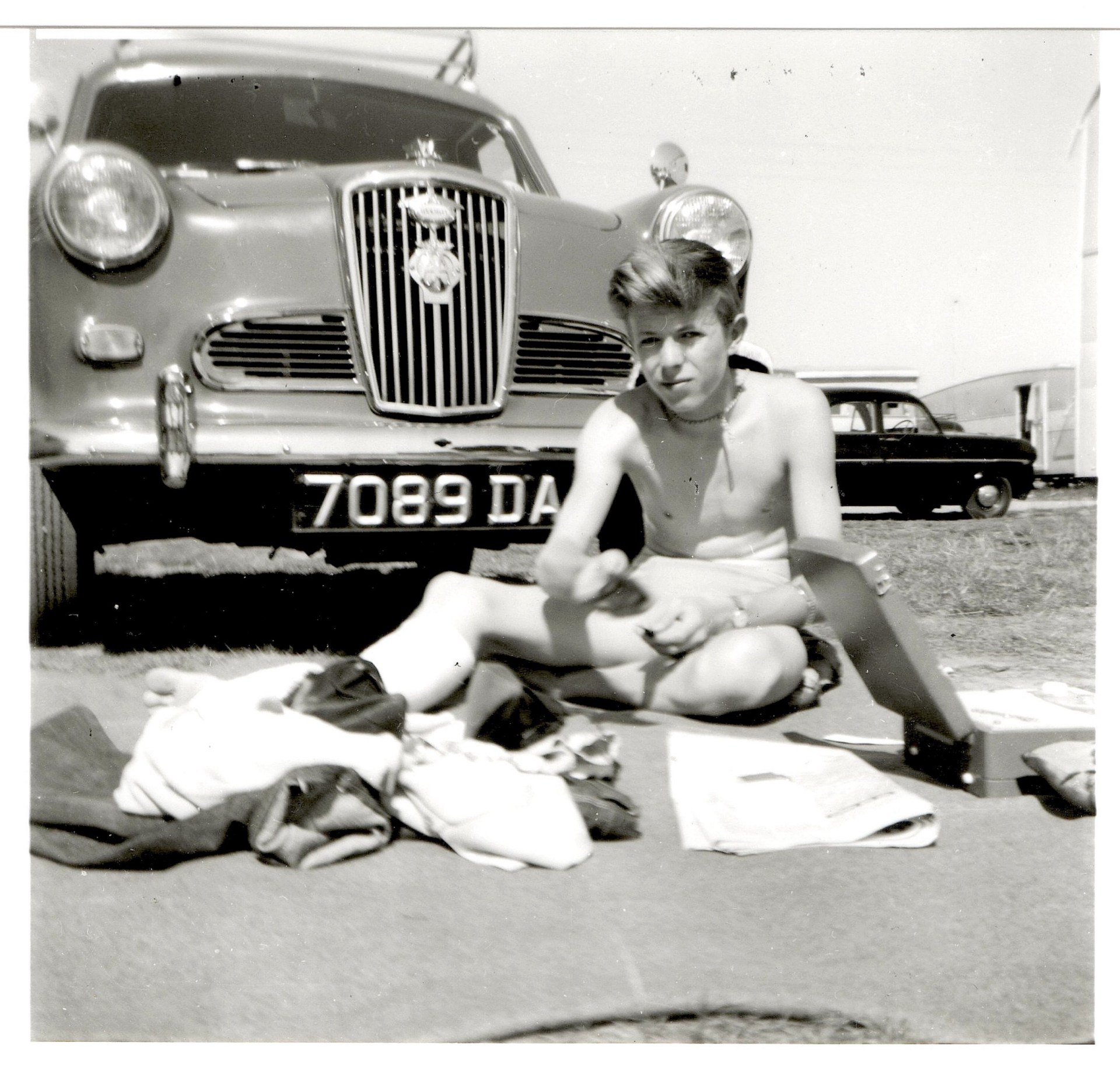 A young David Jones, aka David Bowie, sits in front of a car in a black and white photo