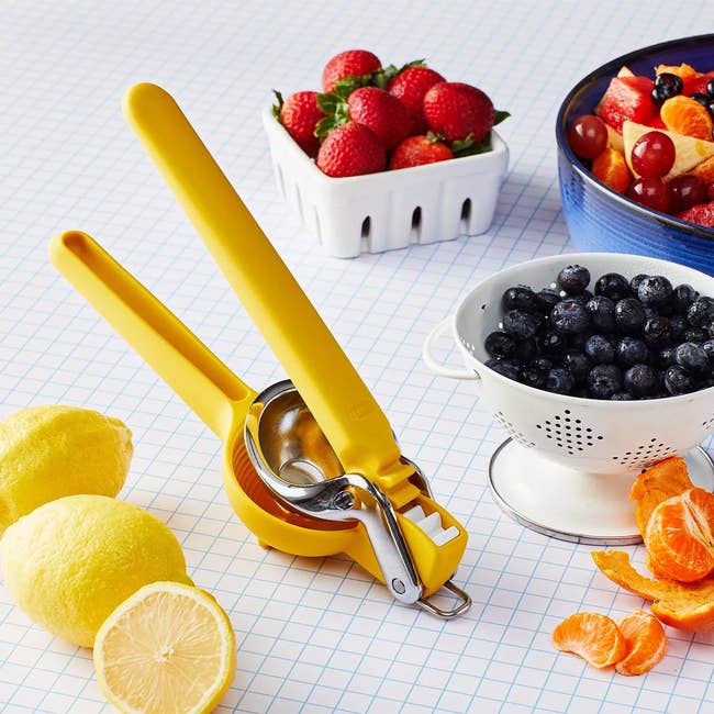 A yellow citrus squeezer is on a table with lemons, berries in bowls, and peeled mandarin segments, suggesting a fresh fruit shopping context