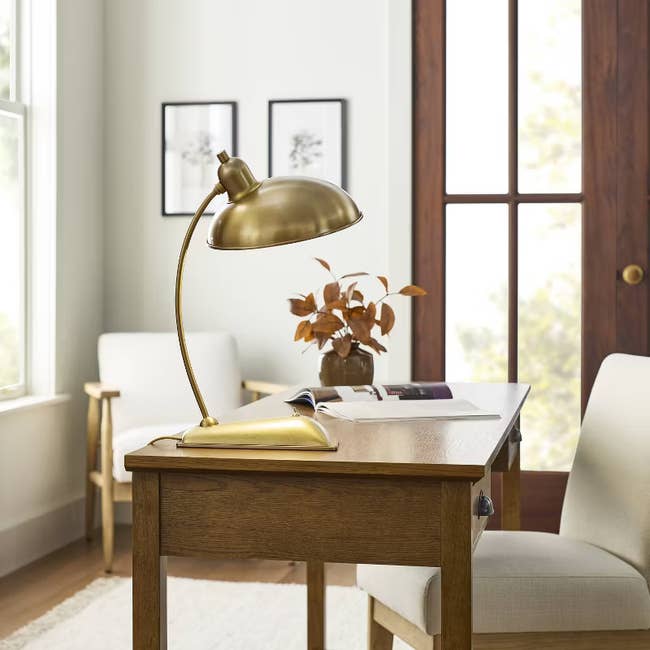 A stylish brass desk lamp on a wooden desk in a modern living space with chairs and framed artwork