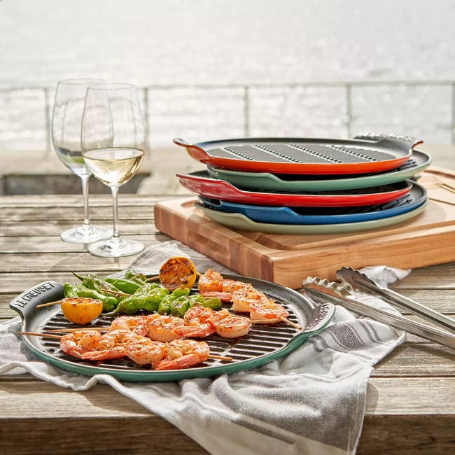 Grill pan with shrimp, roasted lemon, and vegetables on a table outdoors, next to wine glasses and colorful stacked pans