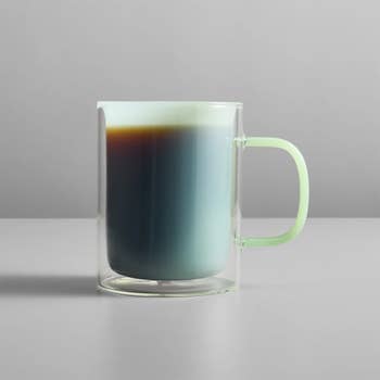 A glass coffee mug with a sleek, minimalist design, featuring a visible coffee layer separation, sits on a gray surface