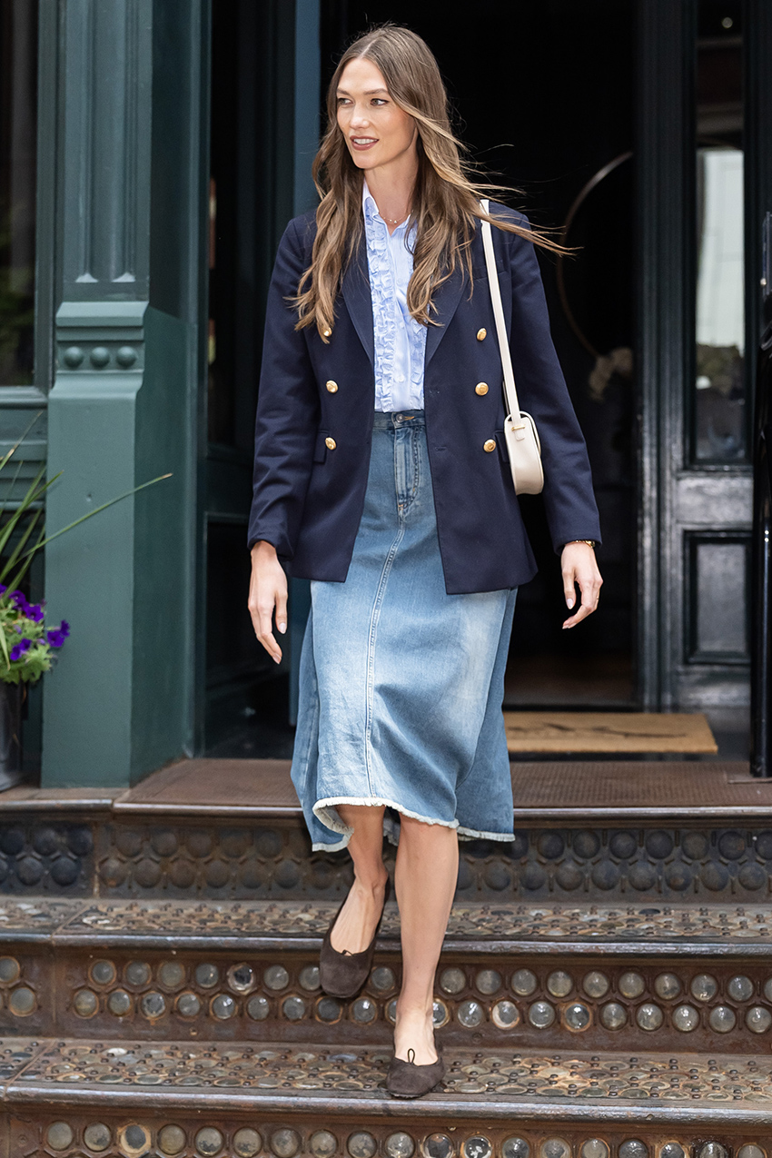 Karlie Kloss wearing a navy blazer over a ruffled blue button-down skirt with a frayed-hem denim midi skirt and brown suede ballet flats.