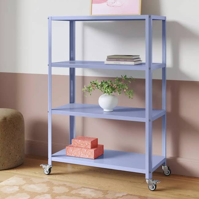 Mobile shelving unit with four tiers, featuring books, a plant in a vase, and decorative boxes