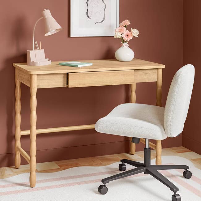 Wooden desk with a drawer, white office chair, lamp, book, and vase with flowers on top; artwork hangs on the wall