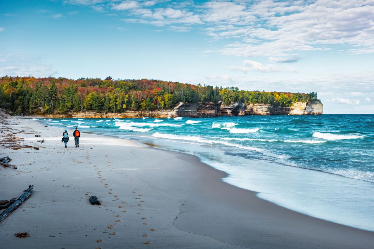 Two people walking on a beach with scenic waves and a forested cliff in the background