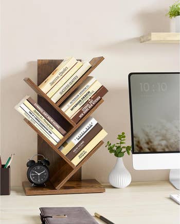 Desktop with zigzag wooden bookshelf holding books, a black alarm clock, and a computer. Minimalist decor with a small vase and notebook