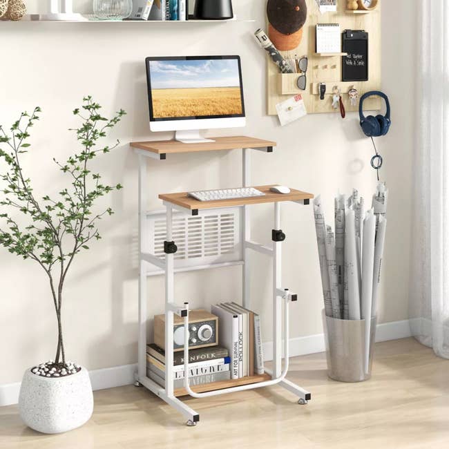 Compact standing desk with monitor, keyboard, and organized workspace accessories. Decor includes a potted plant and rolled-up papers. Ideal for home office