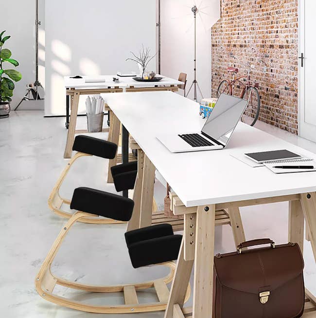 Modern office space with laptops on white desks, ergonomic rocking chairs, and a briefcase on the floor. Brick wall and decor in the background