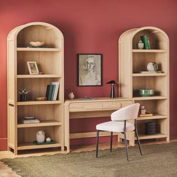 Arched wooden bookshelves with decor flank a wooden desk and chair, set against a red wall with a framed portrait. Plush rug on wooden floor