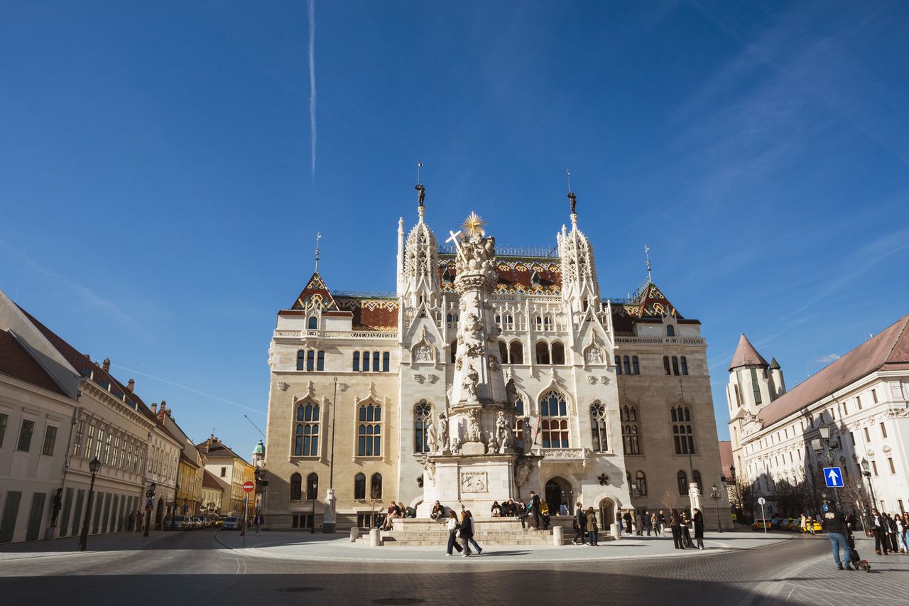 A plaza with a large ornate building and a monument in front people walking and congregating in the square