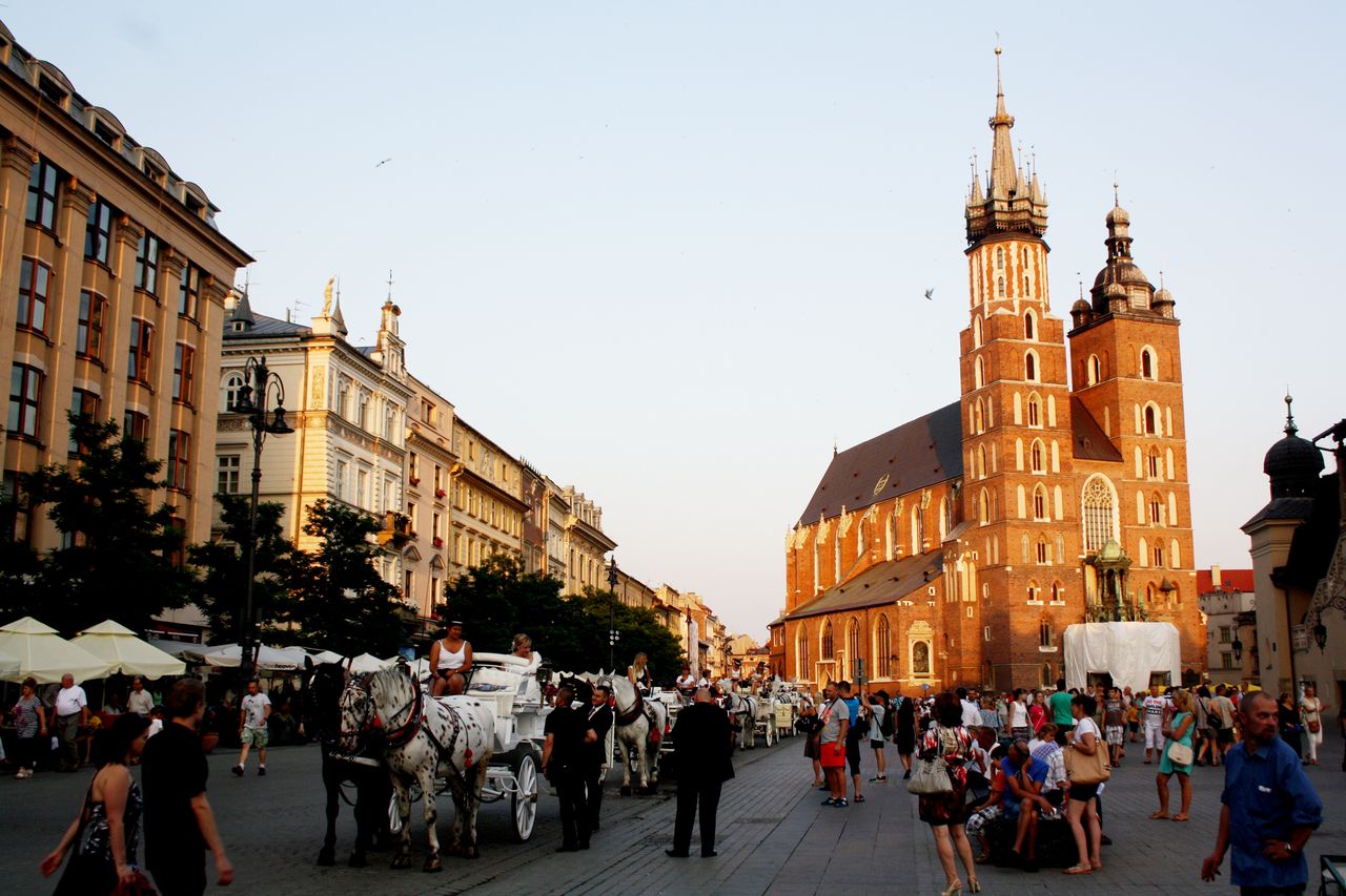 A cobblestone city square featuring a horsedrawn carriage and a large cathedral surrounded by pedestrians and buildings