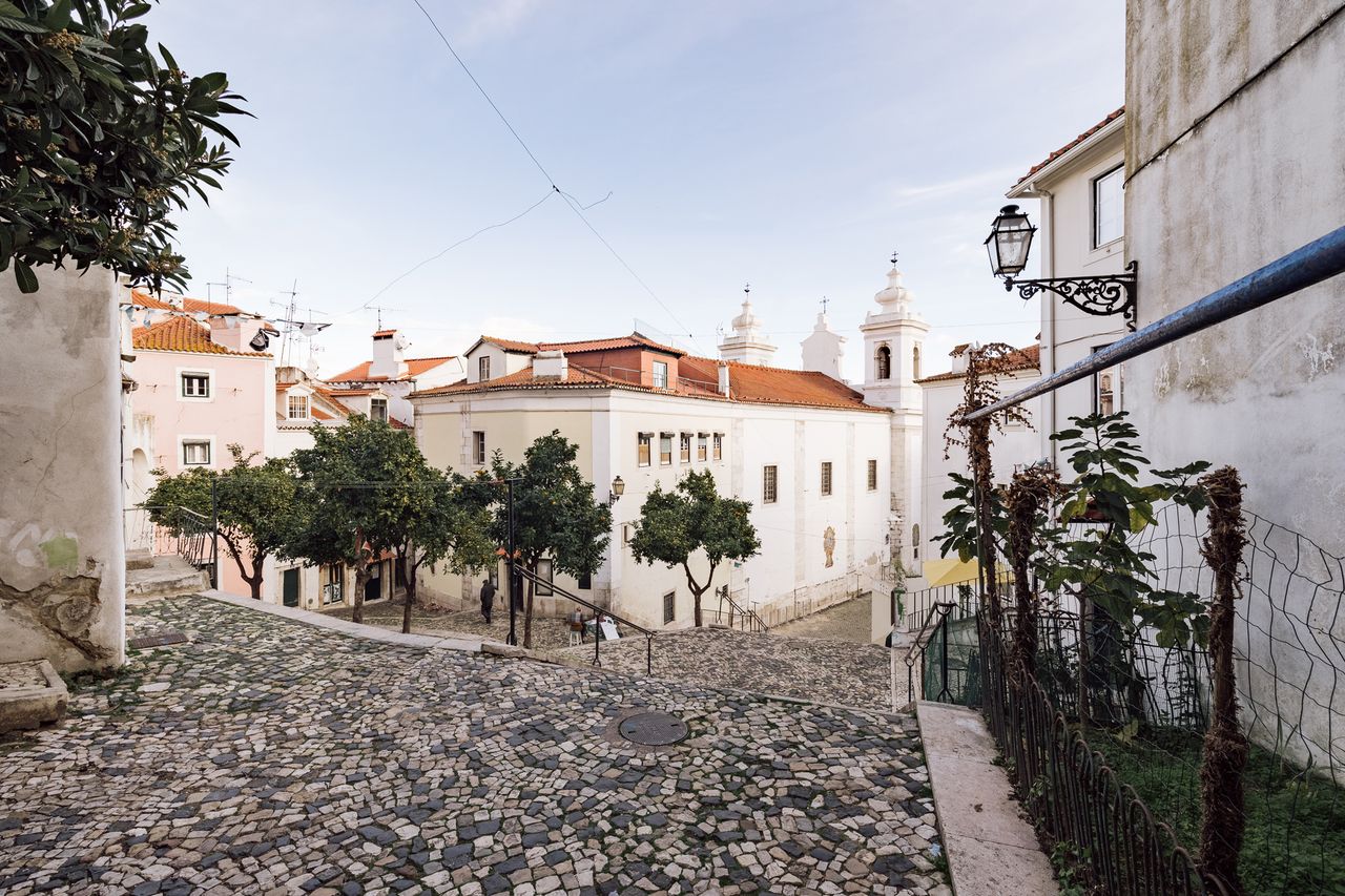 A cobblestone street in a European city with historic buildings and a church in the background surrounded by a few trees and a fenced area