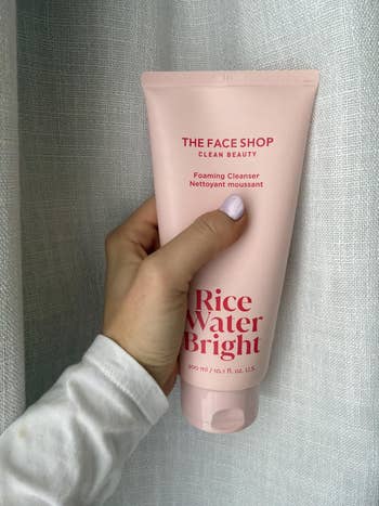 Hand holding a tube of The Face Shop Rice Water Bright Foaming Cleanser against a neutral background