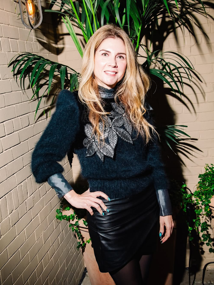 A person posing near a decorated wall with plants wearing a black outfit with floral embellishments