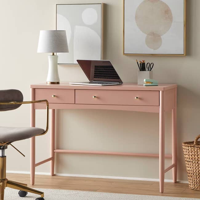 Chic home office desk setup with a pink desk, laptop, lamp, and decor frames. Neutral chair and basket beside. Ideal for stylish, functional workspaces