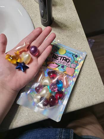Hand holding colorful bath pearls, including star and heart shapes, next to a packet labeled