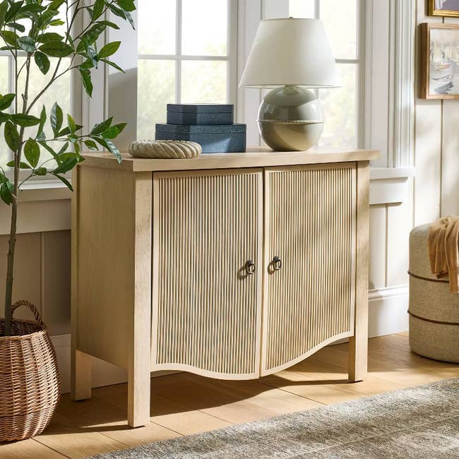 Wooden sideboard in a sunlit room, topped with books, a vase, and a lamp. Woven basket with plant beside it. Cozy, organized decor