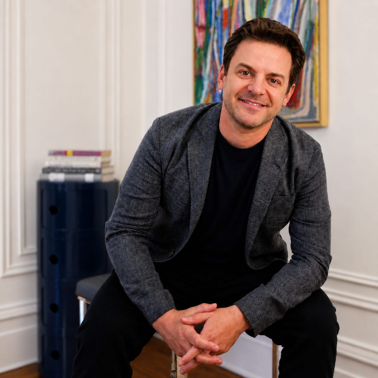 A man in a gray blazer, black shirt, and black pants, sitting on a chair and smiling at the camera.