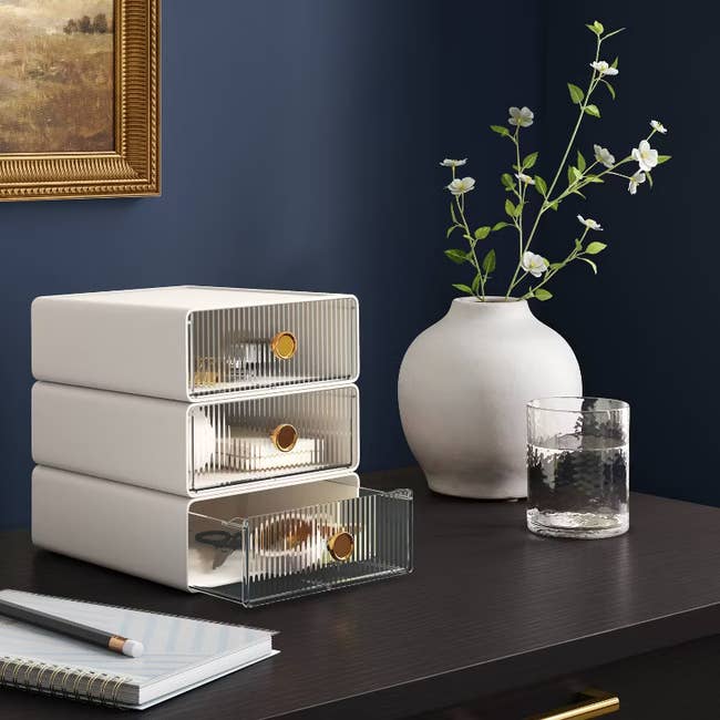 Stacked storage drawers next to a vase with flowers and a glass of water on a dark wood desk