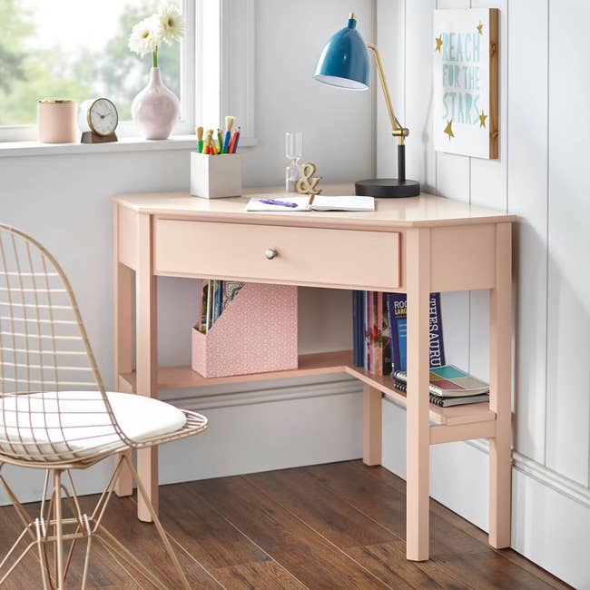 Compact pink desk with drawer and shelves, holding various desk supplies and books, positioned by a window with a lamp and inspirational wall art