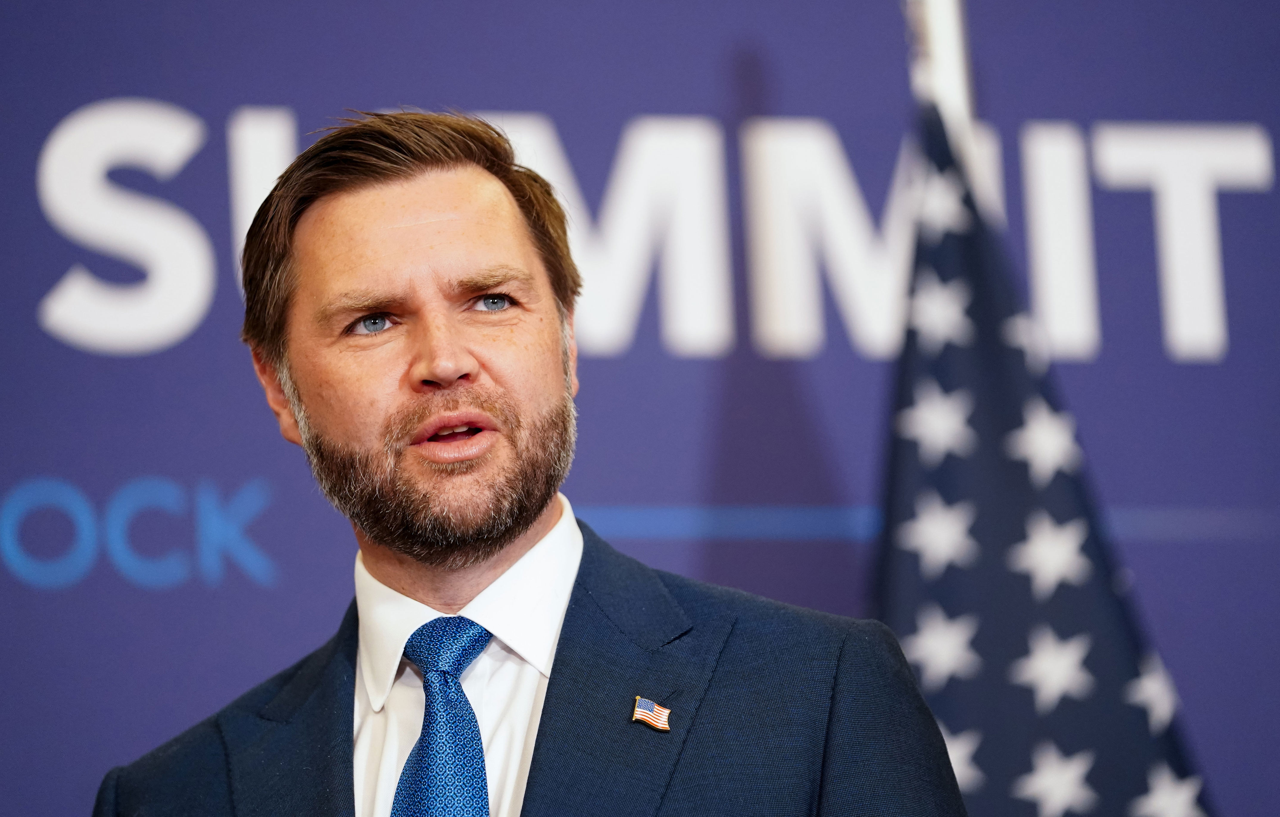 A person in a formal suit speaks at an event with an American flag in the background